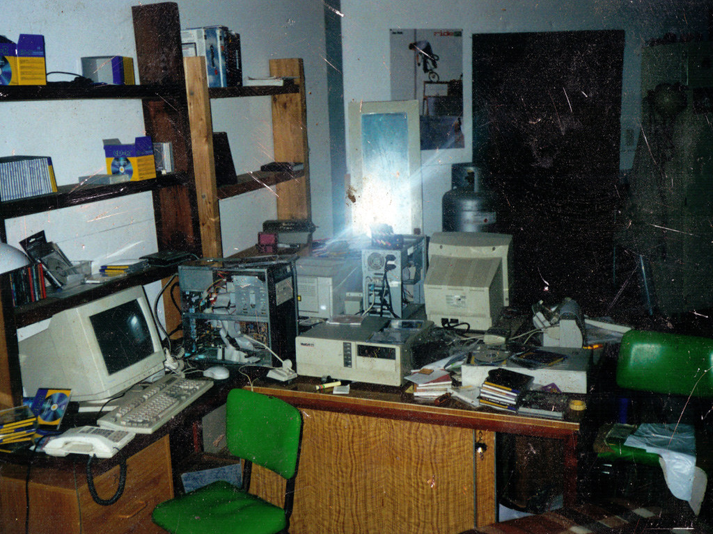 A photo of a very messy set of desks in a garage studio. There are several old 1990s biege-box PCs visible, with loads of burned CD-Rs present. Because it was not very long after the 1980s, there is also a lot of woodgrain and cheap vinyl furniture present.